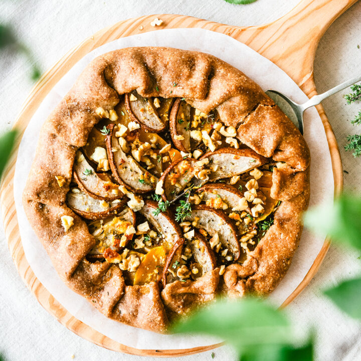 A crispy pumpkin & pear galette placed on a wooden board, ready to be cut into pieces. Sprinkled with walnuts and looked upon through some green leaves.