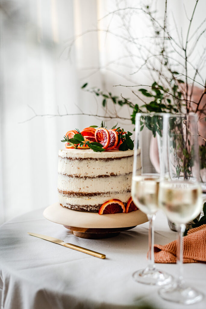 Ein festlich gedeckter Tisch mit einer hellen Schichttorte, getoppt mit Blutorangen und Blättern. Davor, unscharf, zwei Gläser Sekt.