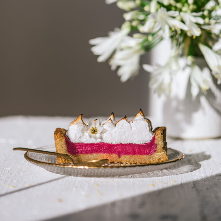 A slice of pink, bright rhubarb pie with toasted meringue placed in front of a white vase with white flowers.