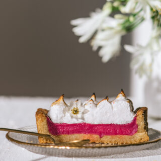 A slice of pink, bright rhubarb pie with toasted meringue placed in front of a white vase with white flowers.