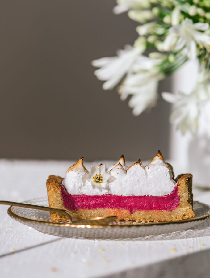 A slice of pink, bright rhubarb pie with toasted meringue placed in front of a white vase with white flowers.