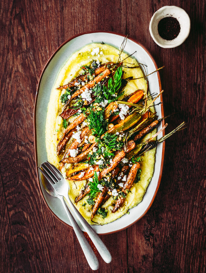 An oval-shaped plate with bright orange roasted carrots layered on top of creamy, yellow polenta & topped with a bright, green carrot-top pesto.