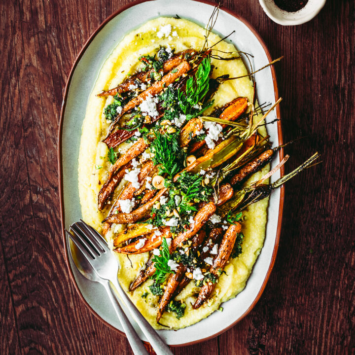 An oval-shaped plate with bright orange roasted carrots layered on top of creamy, yellow polenta & topped with a bright, green carrot-top pesto.