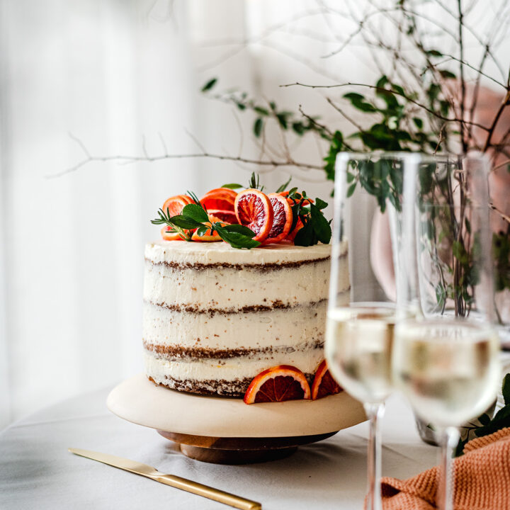 Ein festlich gedeckter Tisch mit einer hellen Schichttorte, getoppt mit Blutorangen und Blättern. Davor, unscharf, zwei Gläser Sekt.